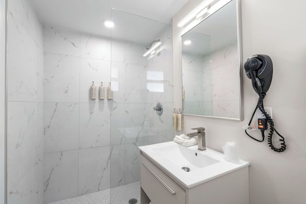A modern bathroom with a marble shower, a white sink, and a hairdryer mounted on the wall. - The Garden Hotel & Resort (Fort Lauderdale)