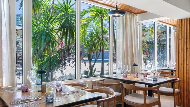 A restaurant with large windows, wooden furniture, and a view of palm trees.