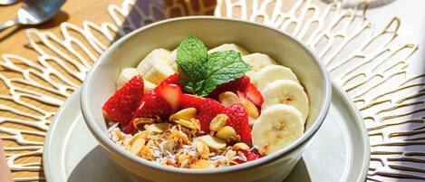 Ein Frühstückstisch mit einer Schüssel Müsli, belegt mit Erdbeeren, Bananen und Nüssen, einem Glas Orangensaft und einem Glas Wasser, dazu eine Tischmatte mit Dekor.
