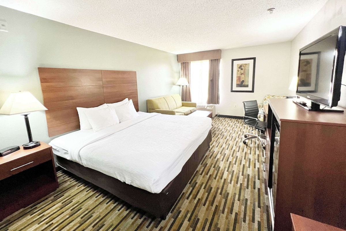 A hotel room with a large bed, a desk with a television, a chair, and a lamp.
