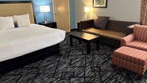A hotel room with a bed, a sofa, a small table, and a chair. There is a door and a lamp on a nightstand. - Baymont by Wyndham Litchfield (Litchfield)