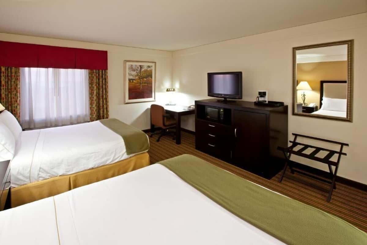 Ett hotellrum med två sängar, ett skrivbord, en tv, en spegel och ett fönster med gardiner.