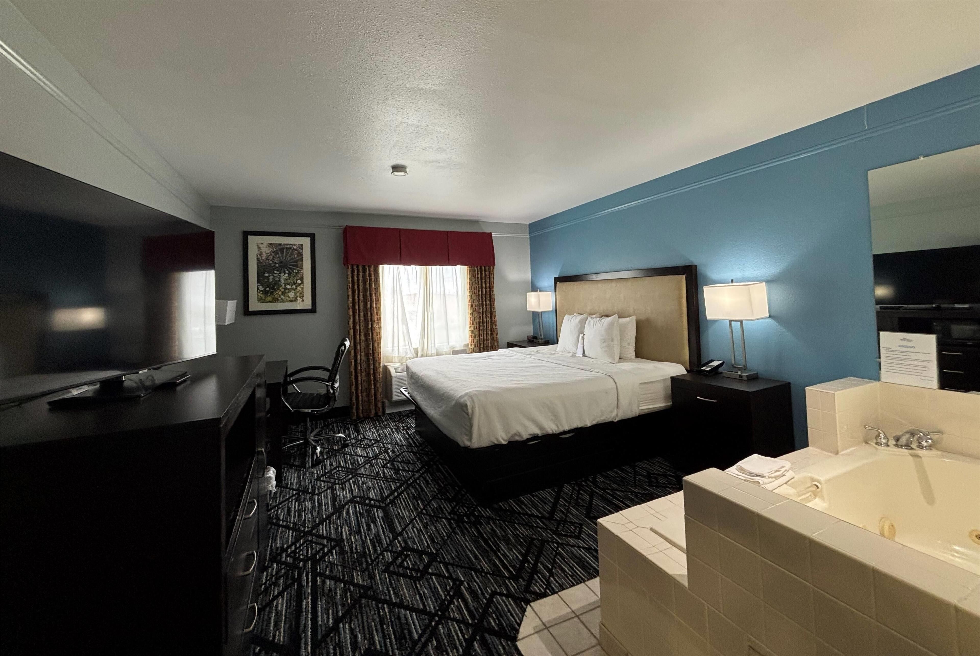 A hotel room with a bed, a desk, a chair, a desk lamp, a TV, a bathroom with a bathtub, and a large mirror.
