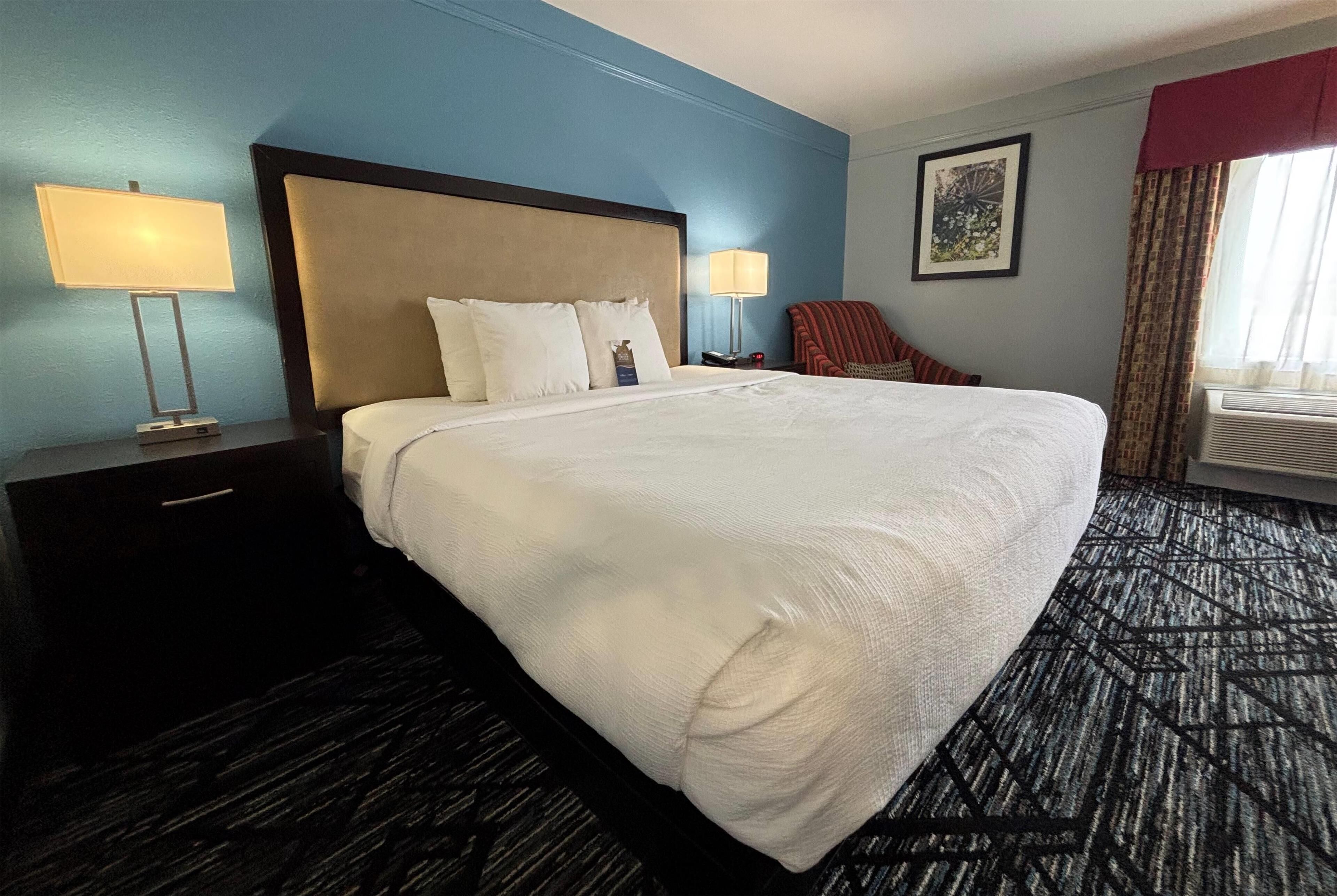 A hotel room with a large bed, two bedside tables with lamps, a chair, a framed picture on the wall, and an air conditioning unit.