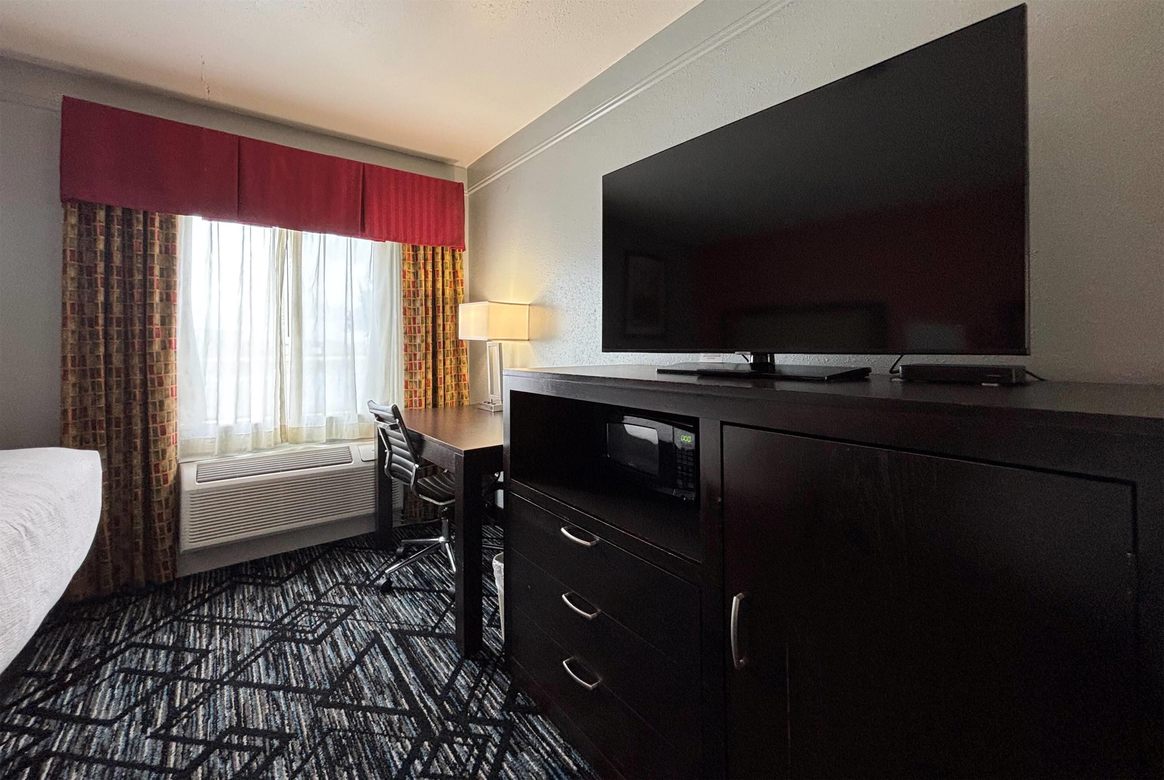 A hotel room with a bed, a desk with a chair, a TV, a microwave, and a large window with curtains.