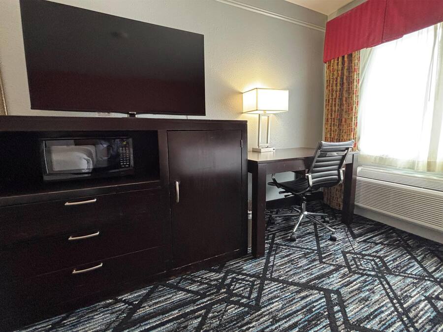 A hotel room with a dark wood cabinet, a microwave, a desk with a chair, a lamp, and a large window with curtains.