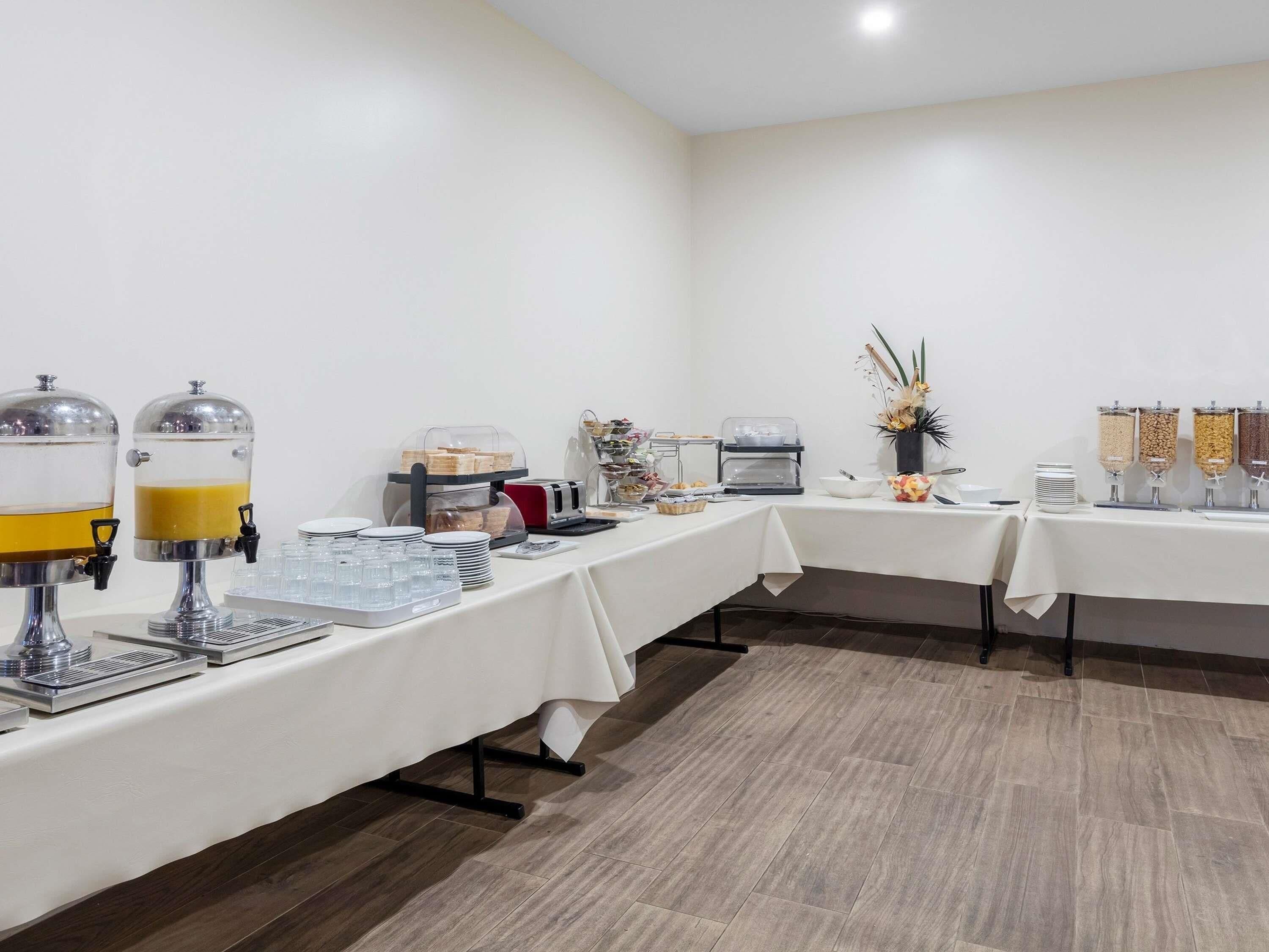 A buffet setup with self-serve drink dispensers, a coffee machine, and various food items on a white tablecloth-covered table.