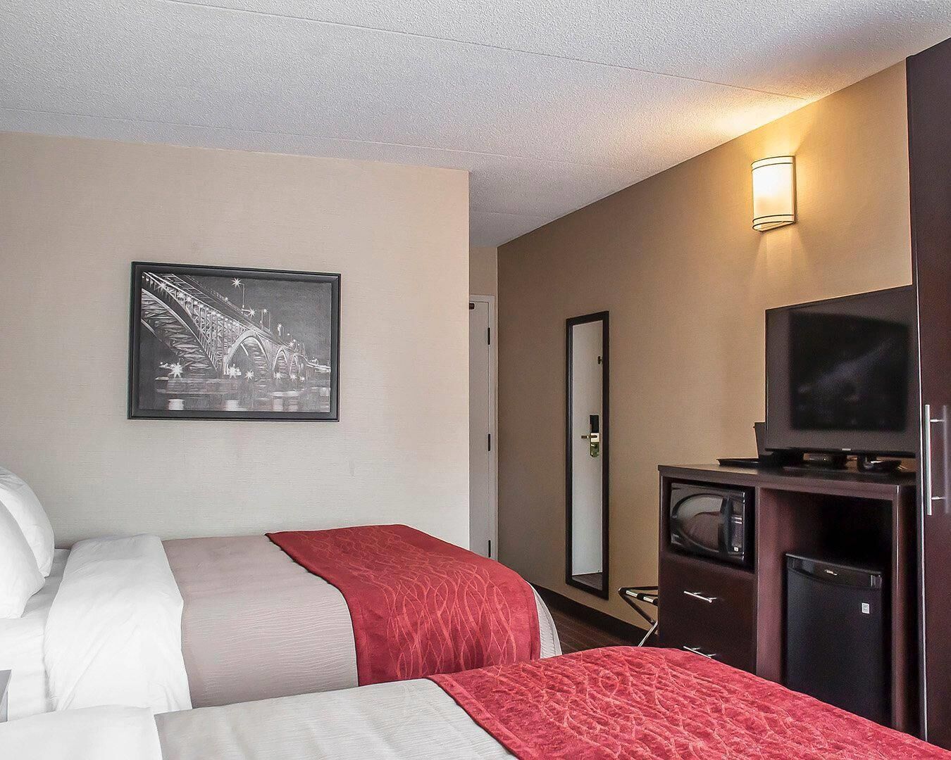 Une chambre d’hôtel avec deux lits, une télévision, un réfrigérateur, un miroir et un tableau encadré au mur.