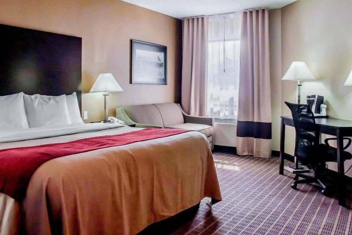 A hotel room with a bed, a desk, a chair, a lamp, a window with curtains, and a framed picture on the wall.