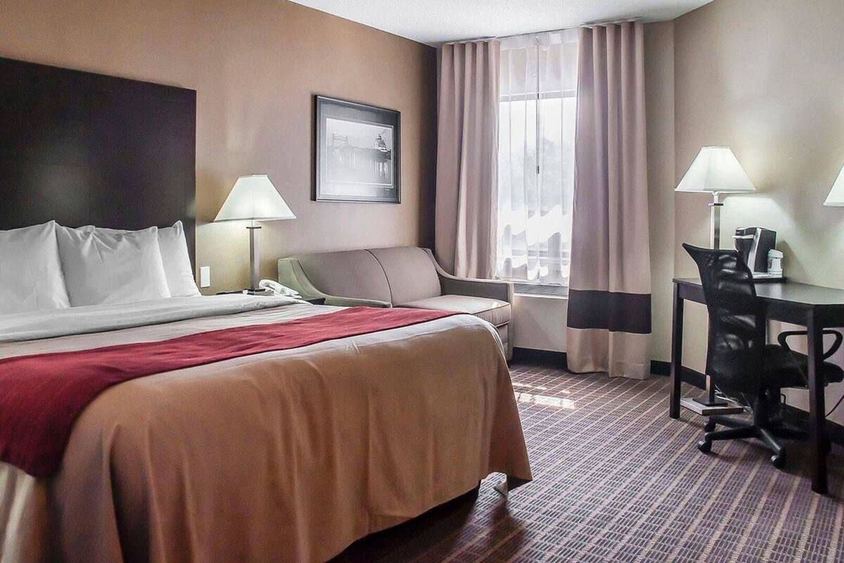 A hotel room with a bed, a desk, a chair, a lamp, a window with curtains, and a framed picture on the wall.