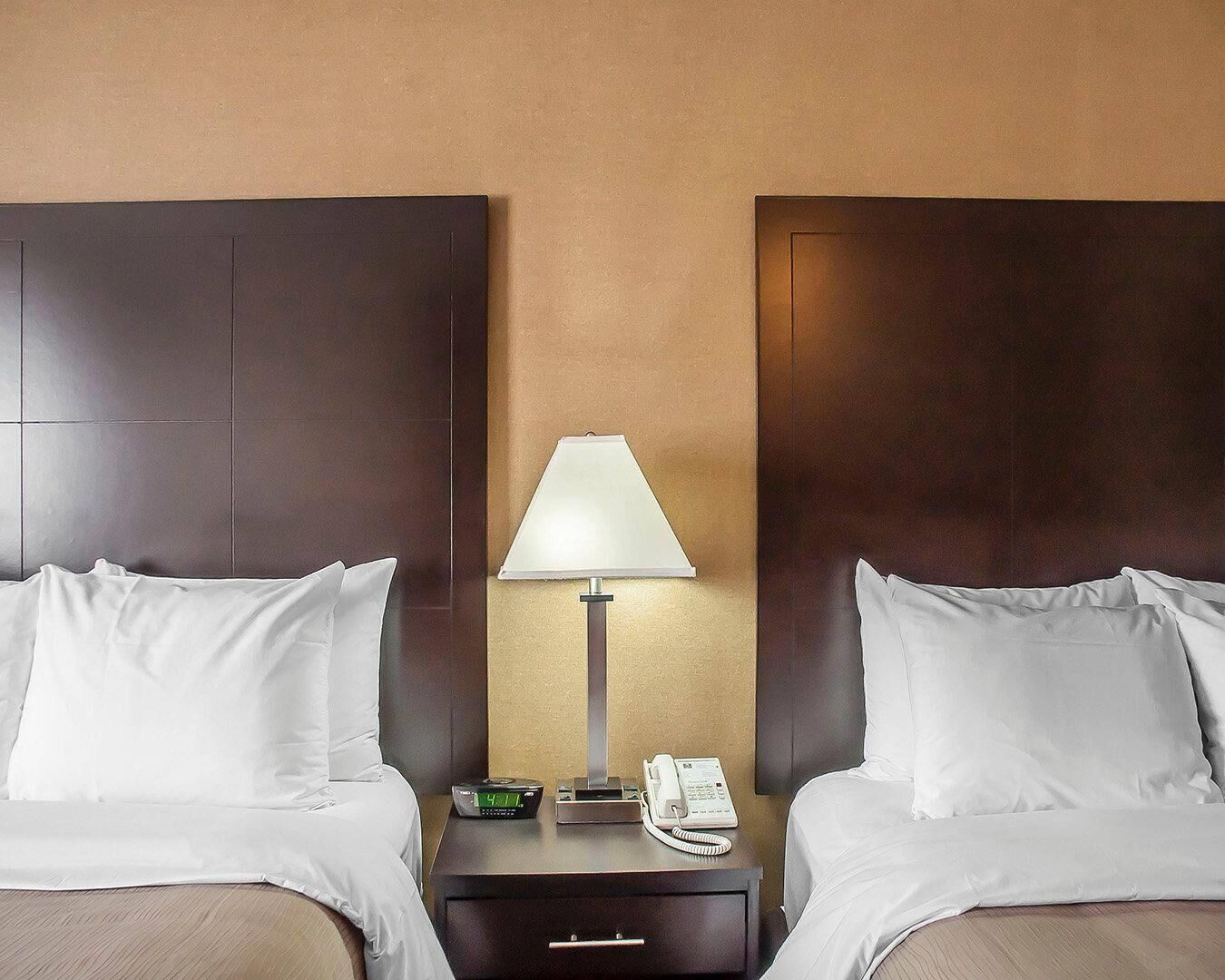 A hotel room with two beds, a nightstand with a lamp, a clock, and a phone.