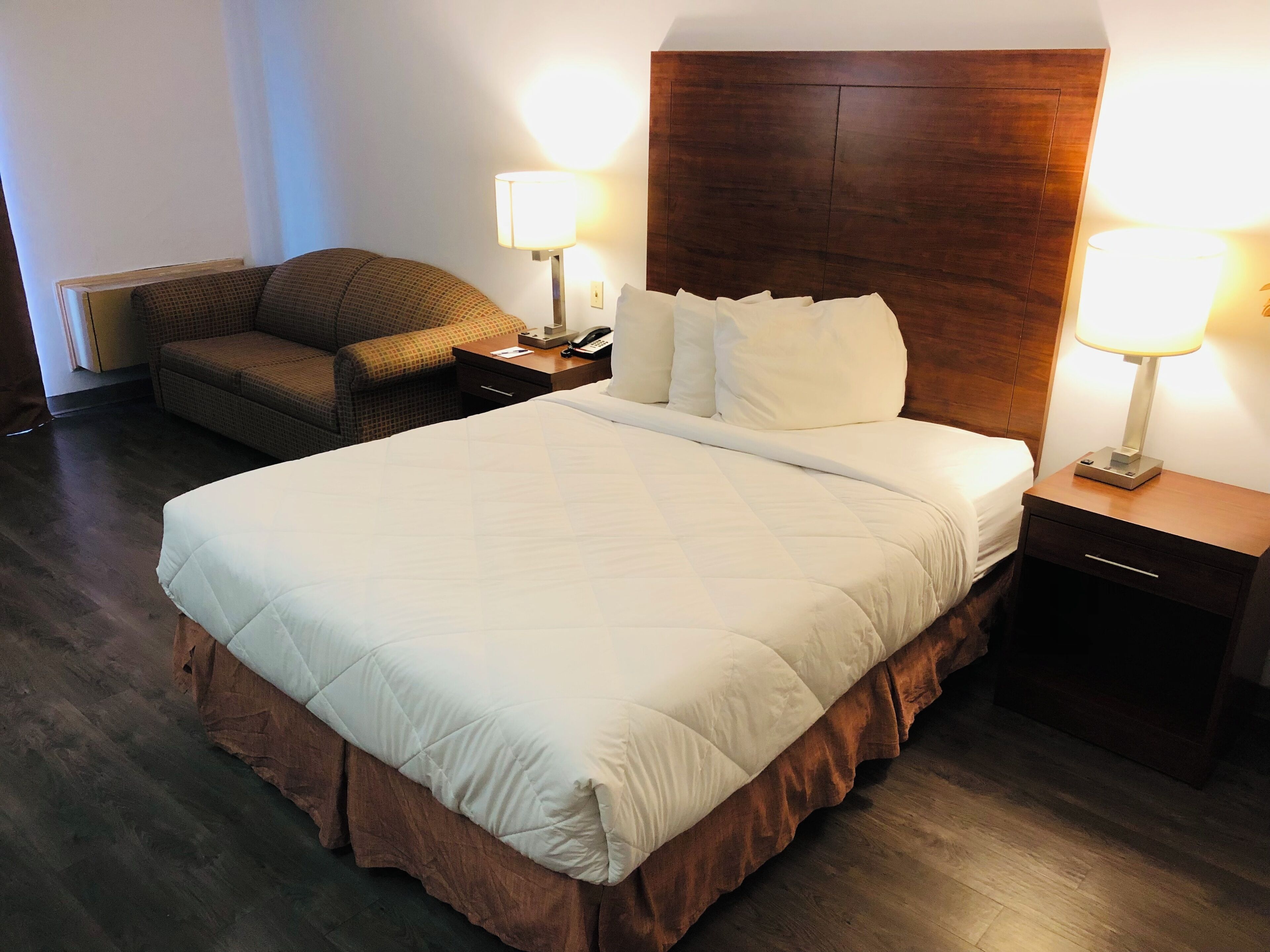 A hotel room with a large bed, a wooden headboard, two bedside tables with lamps, a small sofa, and a nightstand with a phone.