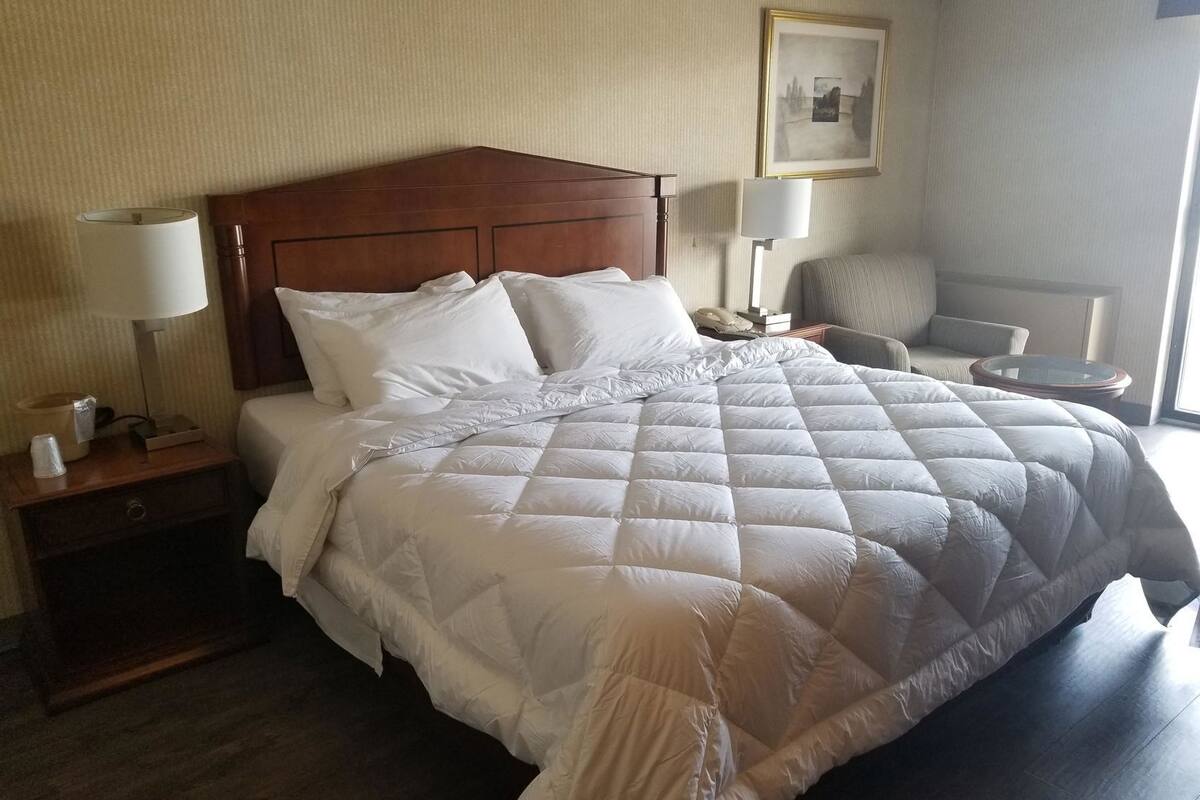 Habitación de hotel con cabecera de madera, cama grande con ropa blanca, dos mesitas de noche con lámparas, una silla e imagen enmarcada en la pared.