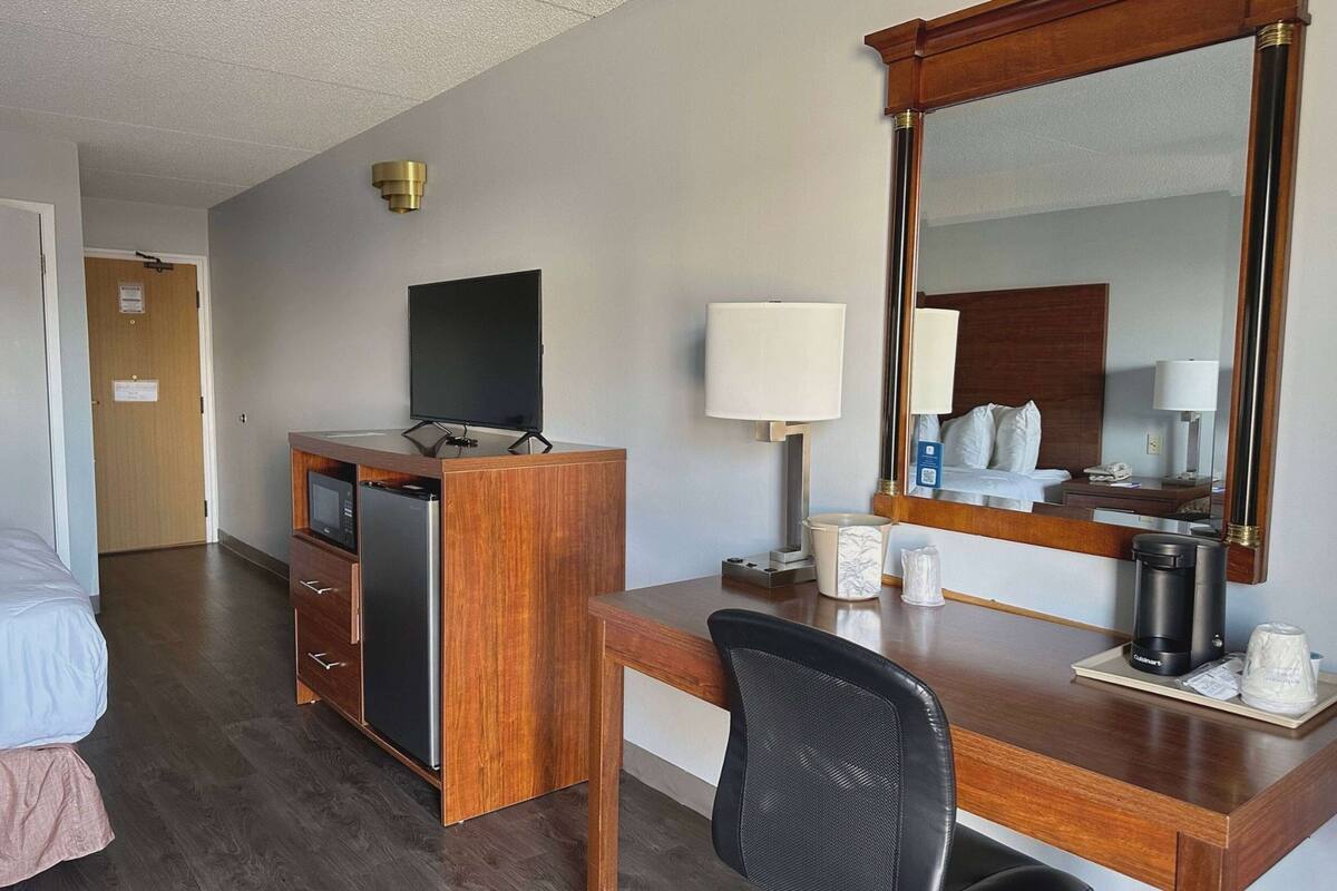 Habitación de hotel con un escritorio de madera, televisor, cama, espejo y cafetera.