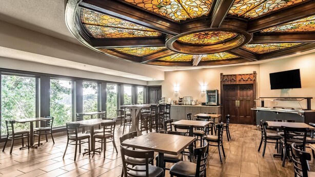 A dining area with wooden tables and chairs, large windows, a kitchen area, and a stained glass ceiling. - Riverview Inn & Suites, an Ascend Collection Hotel (Rockford)