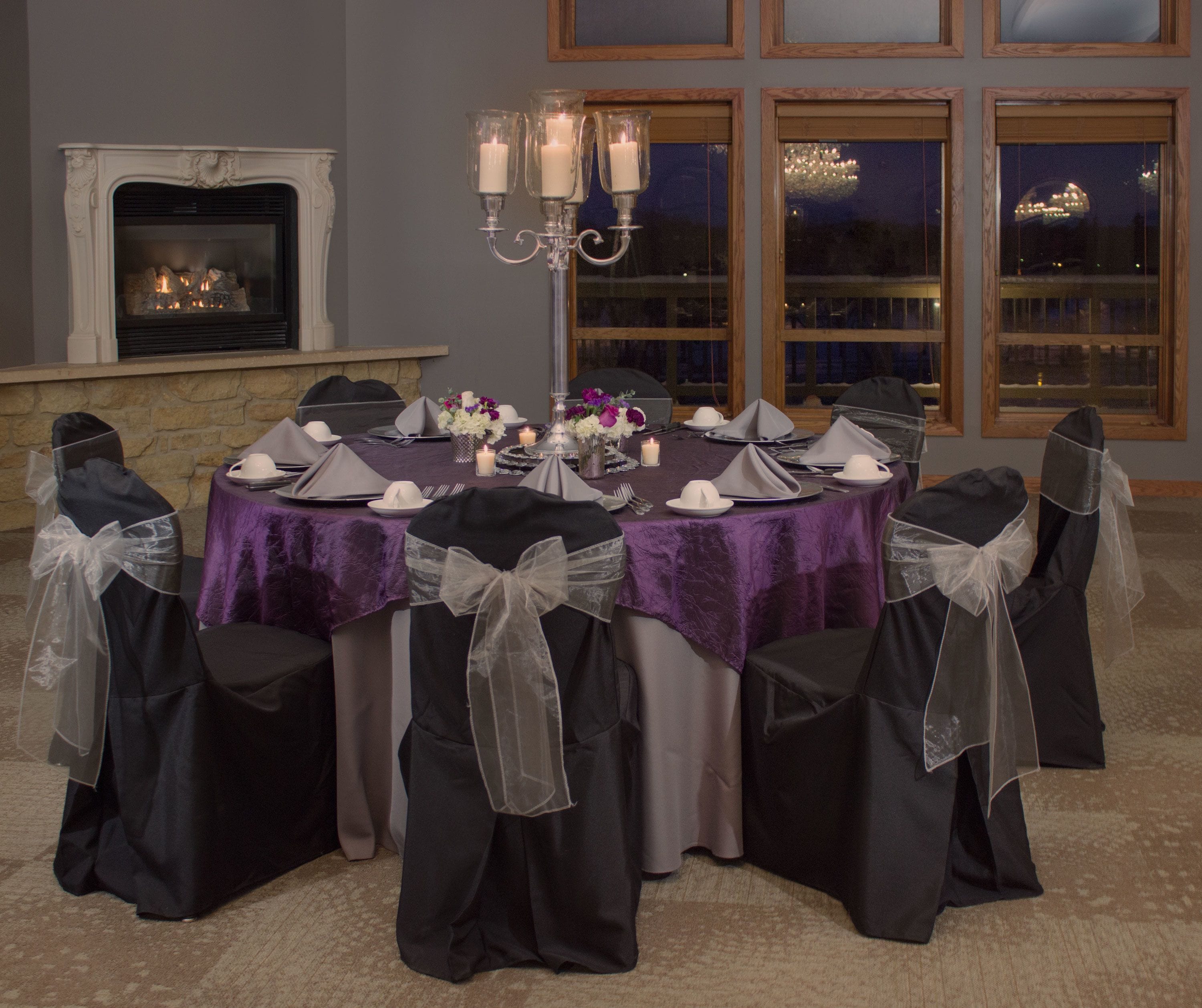 A round table with a purple tablecloth, surrounded by chairs with black covers and white bows.