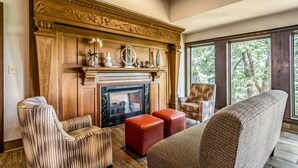 A living room with a fireplace, two red ottomans, a striped armchair, and a large window with a view of trees. - Riverview Inn & Suites, an Ascend Collection Hotel (Rockford)