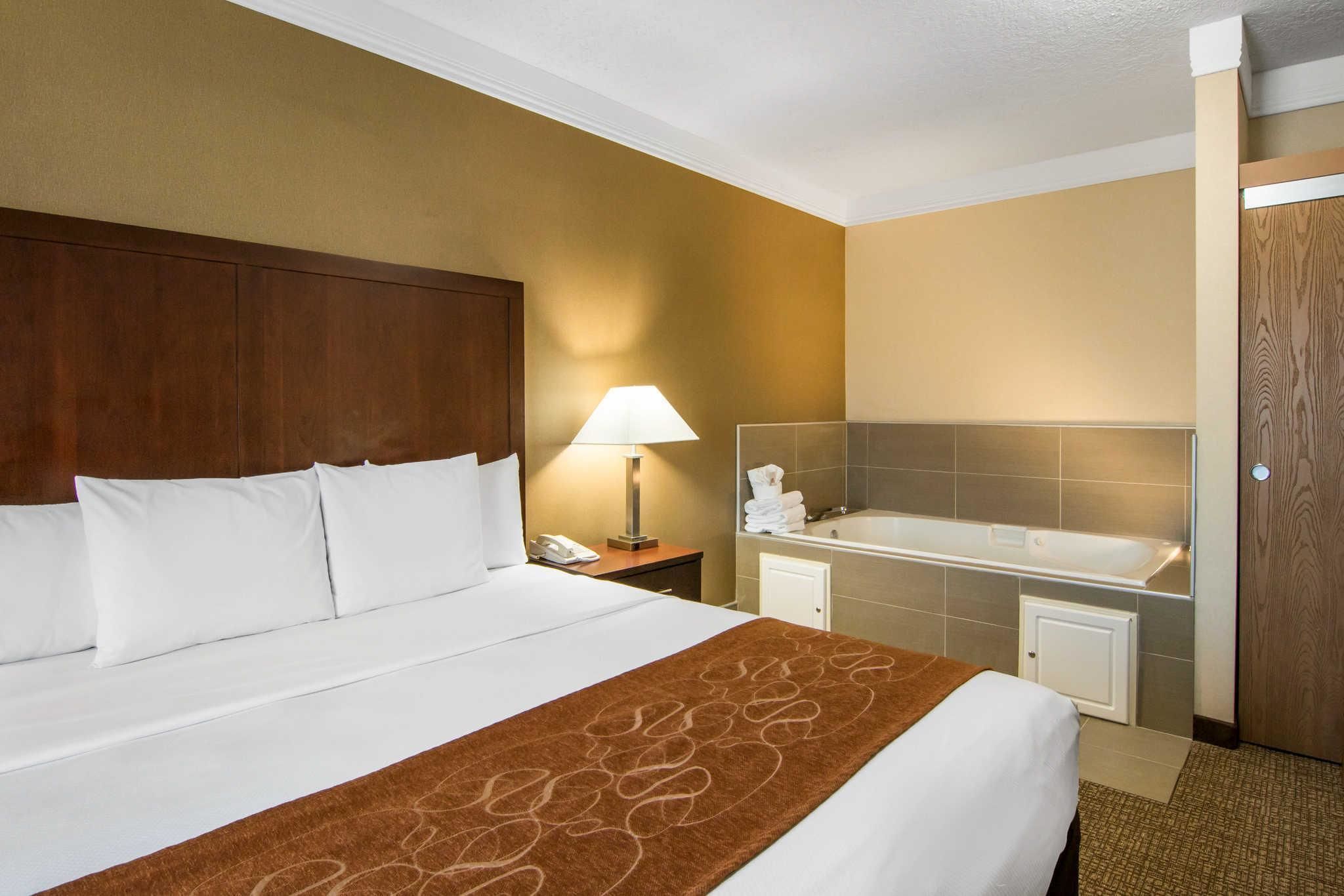 A hotel room with a large bed, a nightstand with a lamp, a bathroom with a bathtub, and a wooden door.