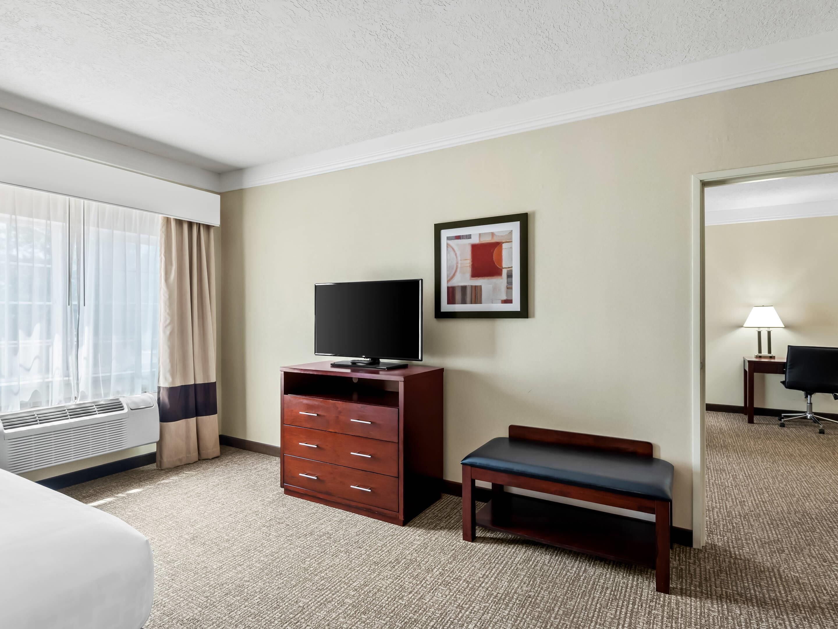 A hotel room with a bed, a TV on a wooden cabinet, a bench, a framed picture on the wall, and a lamp on a side table.