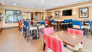 A hotel dining area with multiple tables and chairs, a television mounted on the wall, and a view of the outdoors through large windows. - Comfort Suites Linn County (Albany)
