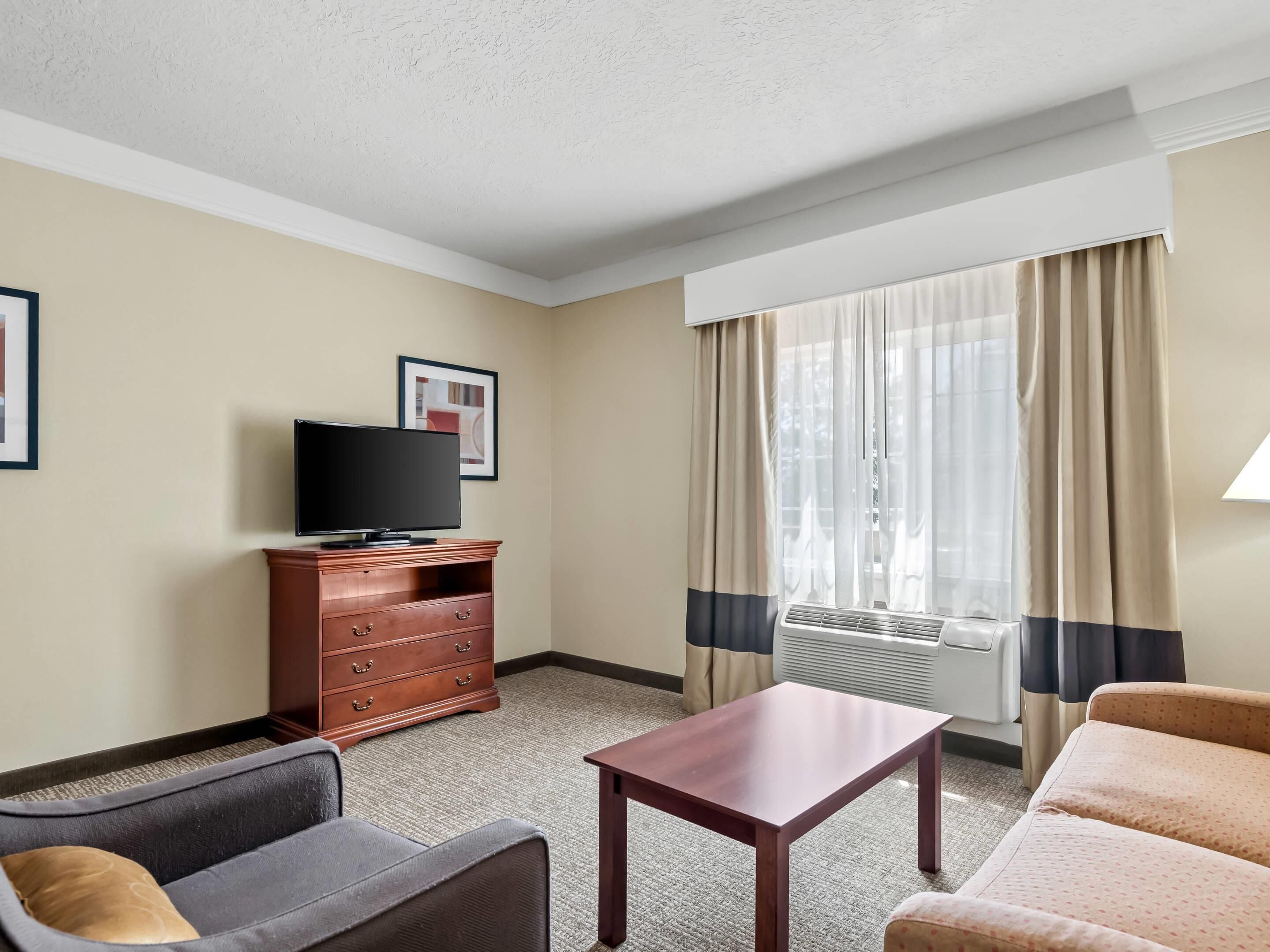 Une chambre d’hôtel avec un téléviseur à écran plat posé sur une commode en bois, un canapé, une petite table et une fenêtre avec des rideaux.