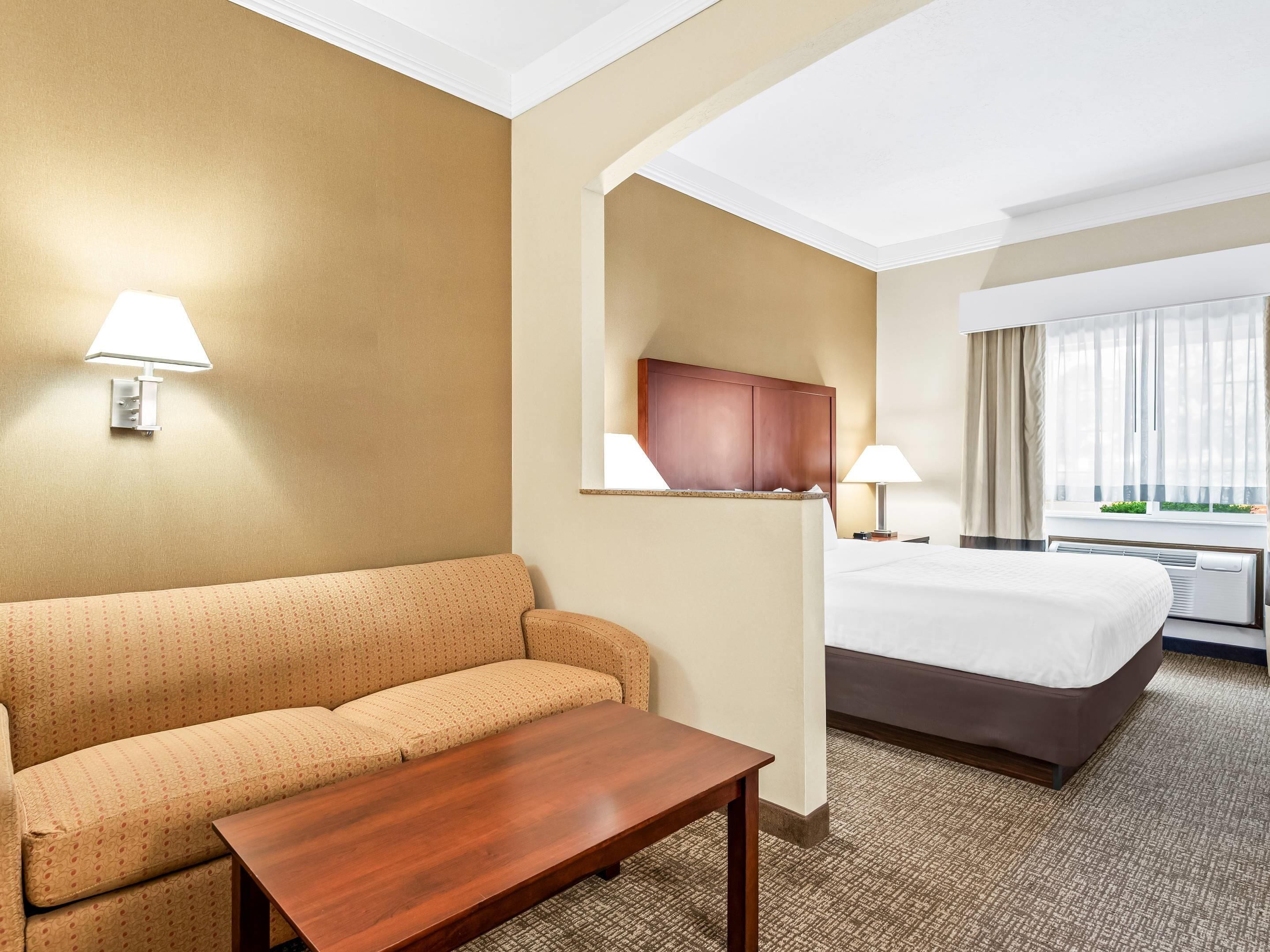 A hotel room with a sofa, a wooden table, a bed, a TV, and a window with curtains.