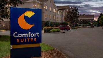 Letrero del hotel Comfort Suites con diseño moderno, un estacionamiento y un edificio al fondo.
