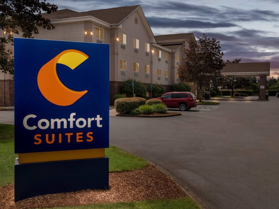 Letrero del hotel Comfort Suites con diseño moderno, un estacionamiento y un edificio al fondo.