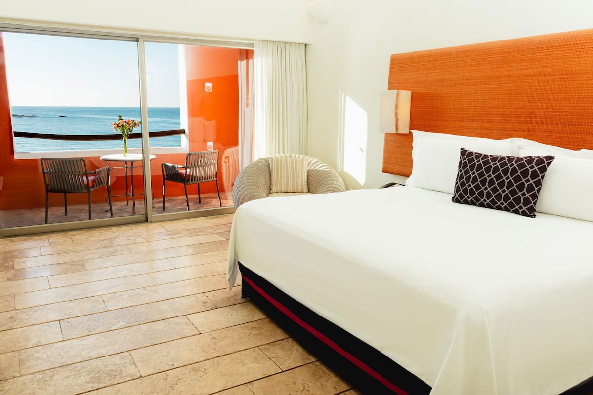 Ein Hotelzimmer mit einem großen Bett, einem Balkon mit Meerblick, einem kleinen Tisch mit Stühlen und einem Fenster mit Vorhängen.