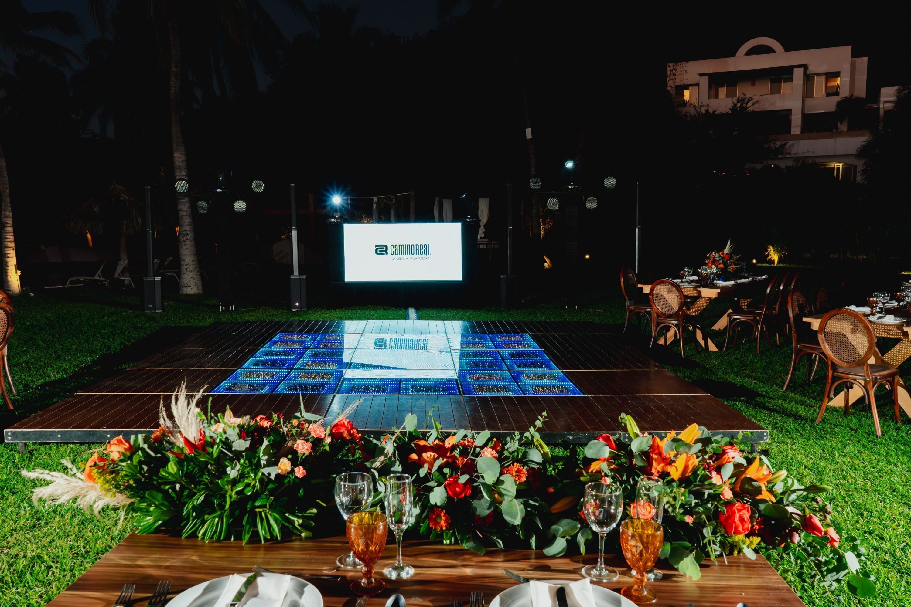 An outdoor dining area with a stage, tables set for a reception, and a large illuminated sign in the background.