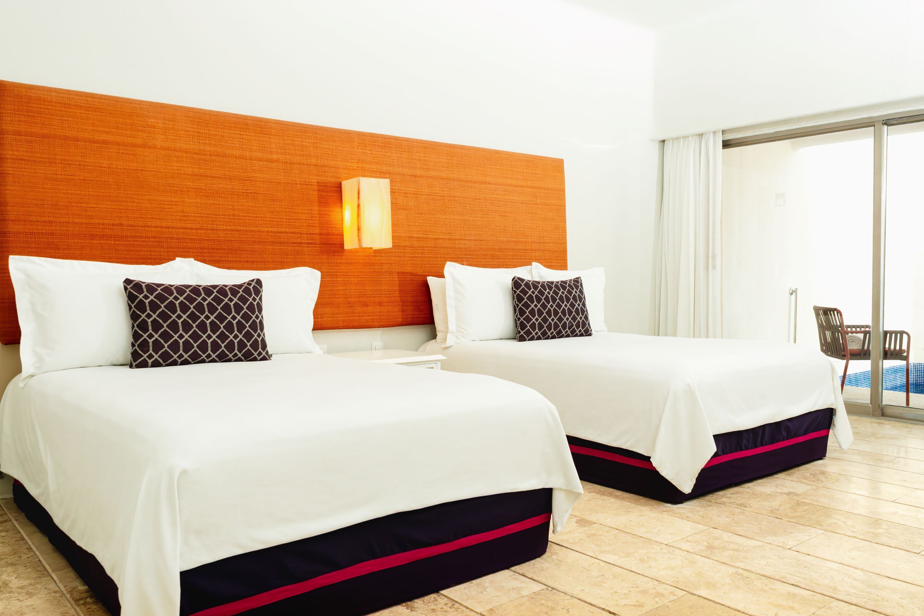 Habitación de hotel con dos camas, cabecero de madera, luz de pared y vista a una piscina a través de un ventanal.