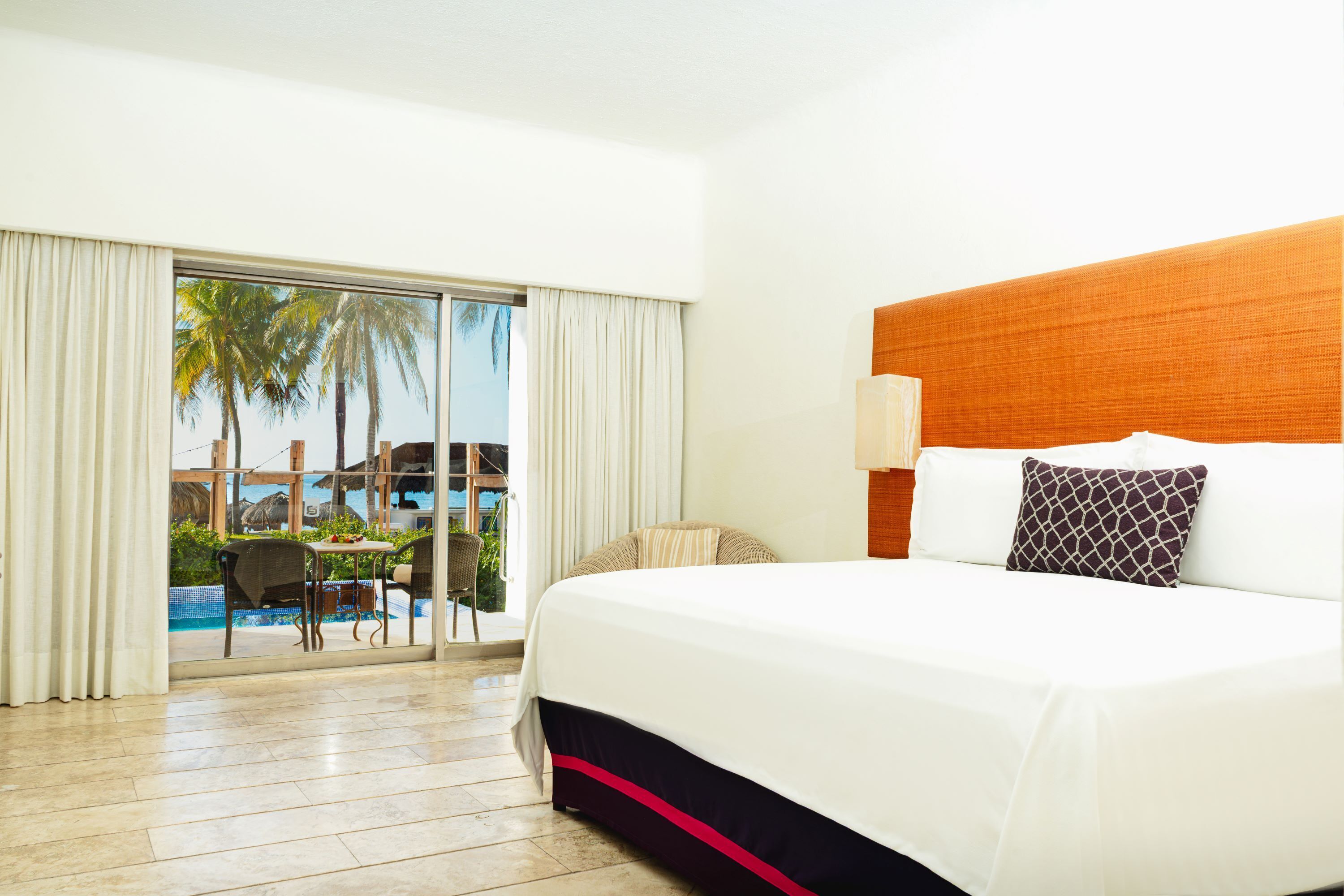 Quarto de hotel com uma cama grande, vista para uma piscina e palmeiras, e área de refeições com mesa e cadeiras.