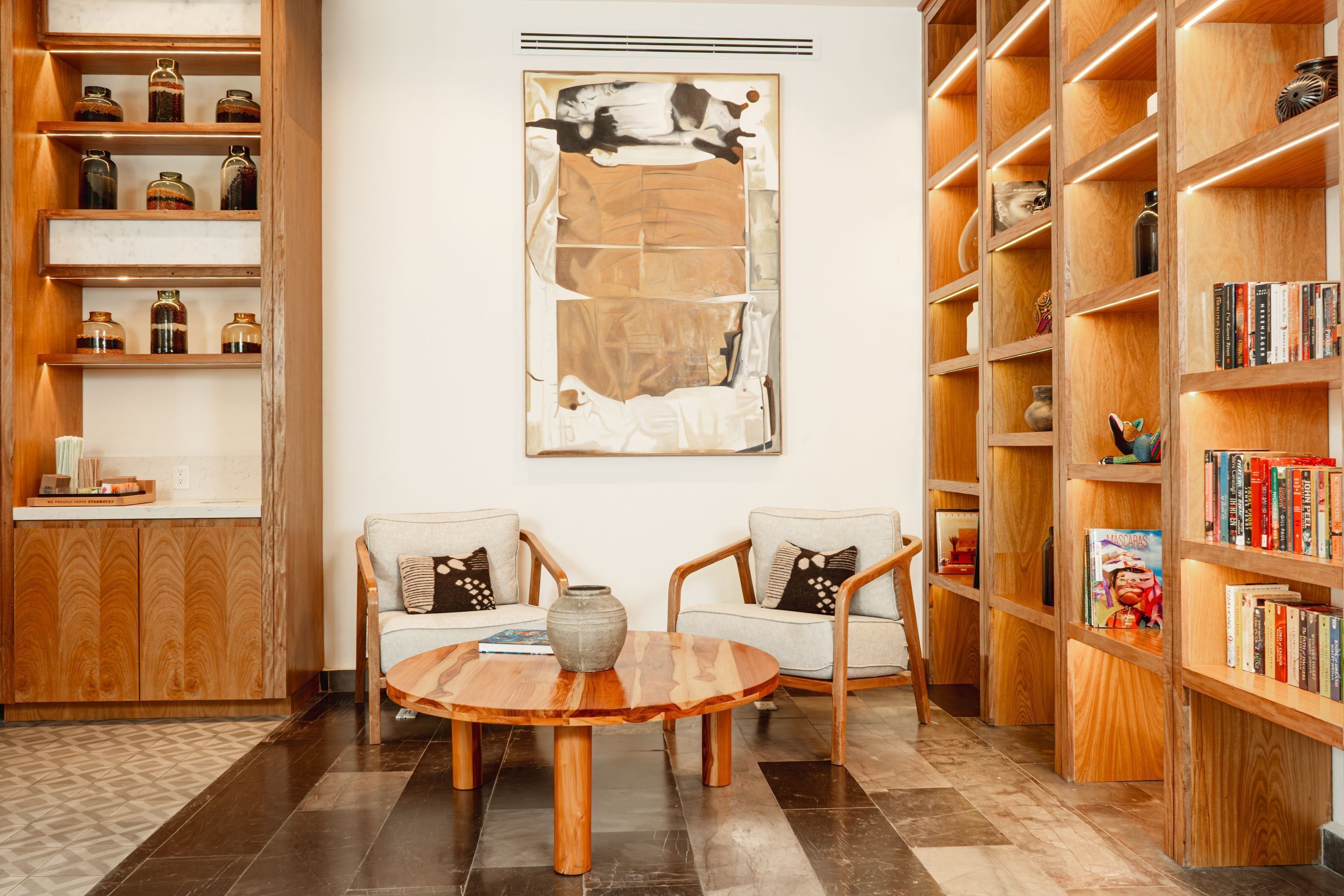 Una biblioteca moderna con estanterías de madera, un cuadro abstracto grande y dos sillones con una mesa de centro redonda de madera.