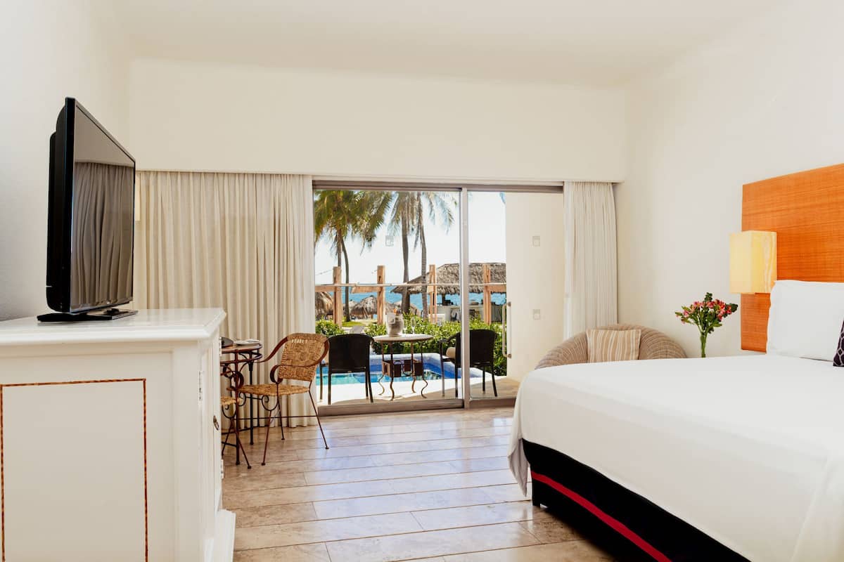 Ein Hotelzimmer mit Bett, Fernseher, Essecke mit Stühlen, Tisch und Blick auf Pool und Palmen.