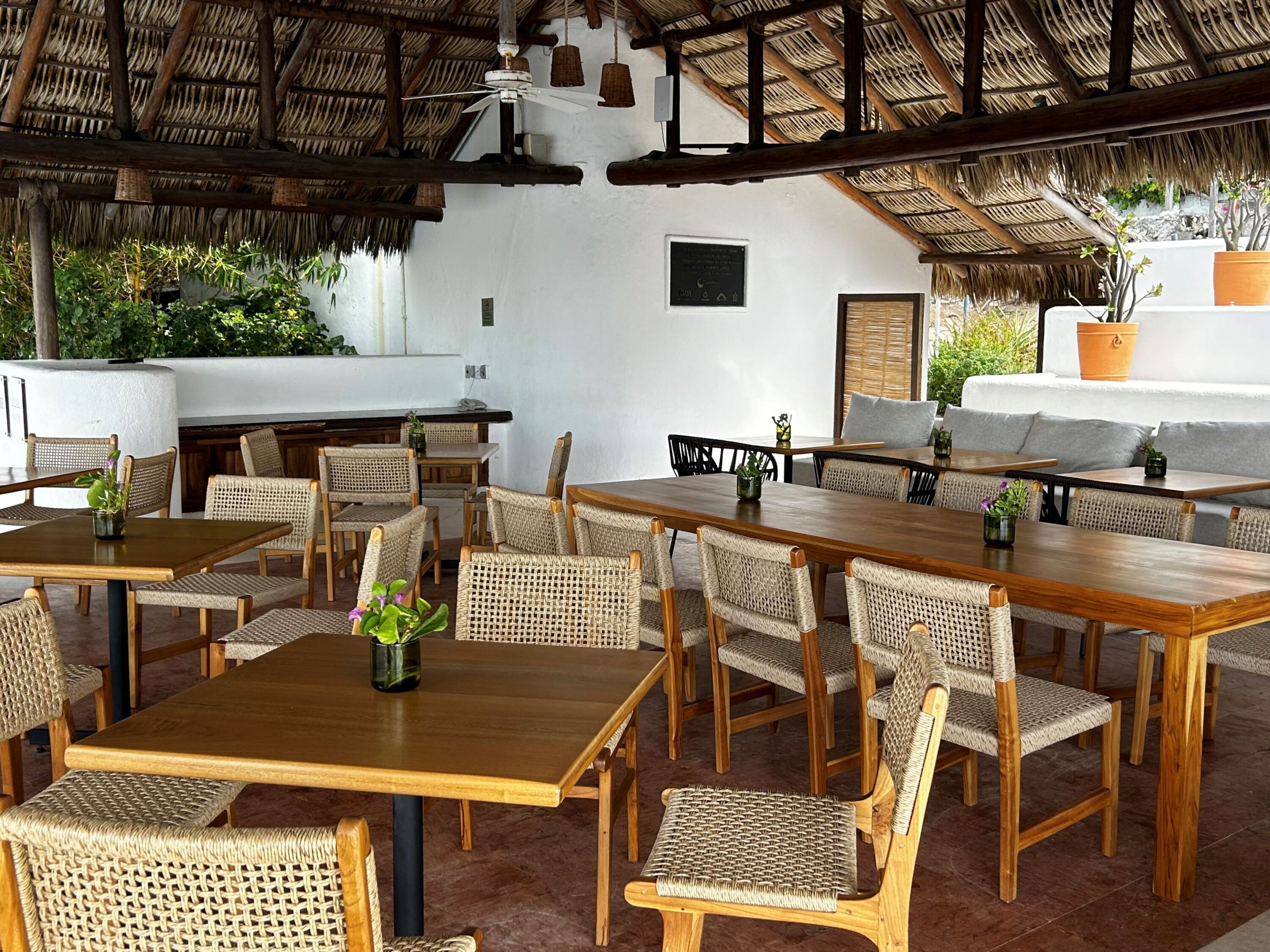 Een restaurant met houten tafels en rieten stoelen, een rieten dak en een witte muur met een ingelijste foto.