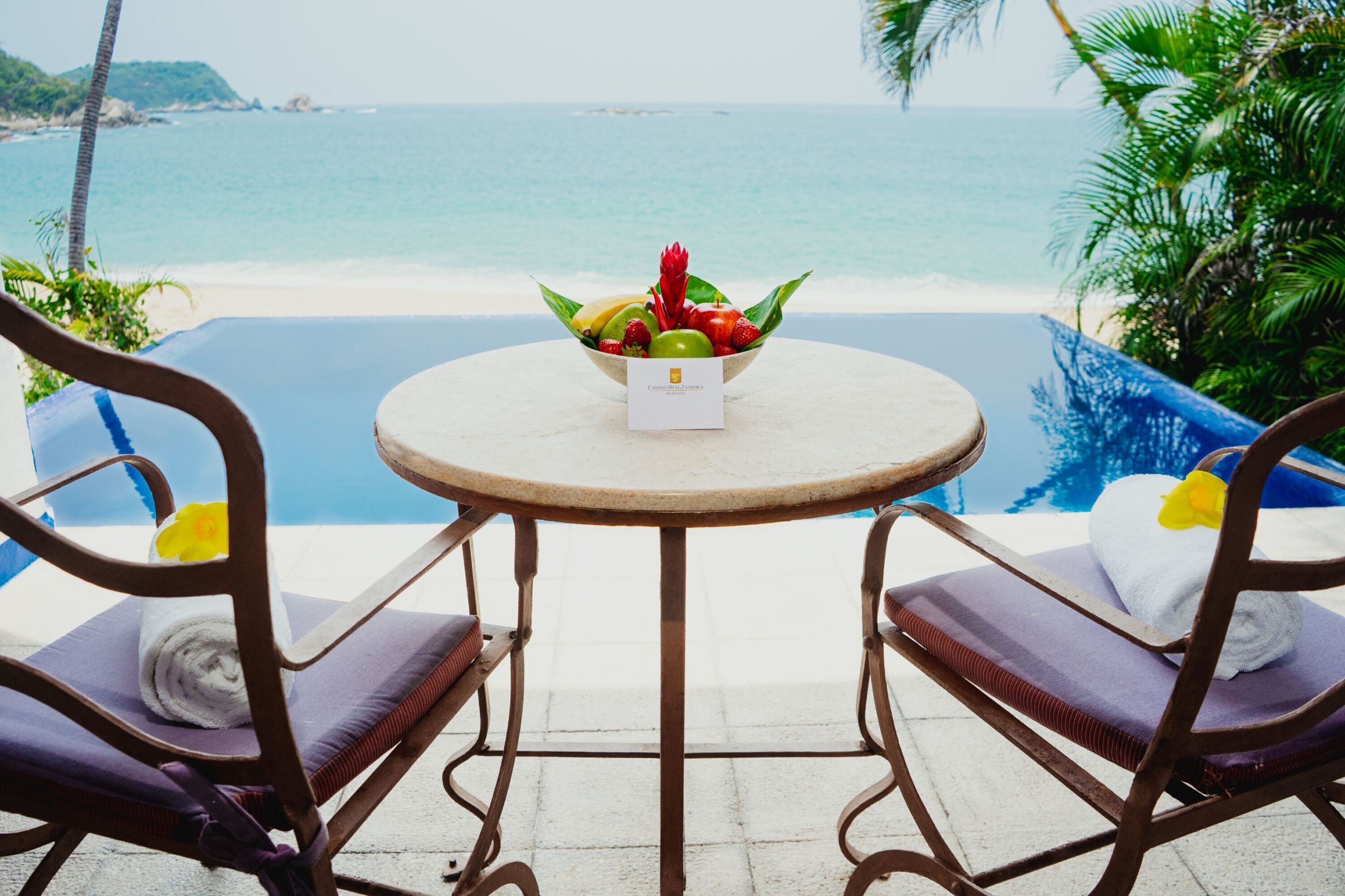 Una mesa con frutero y una tarjeta, con vista a la playa y la piscina.