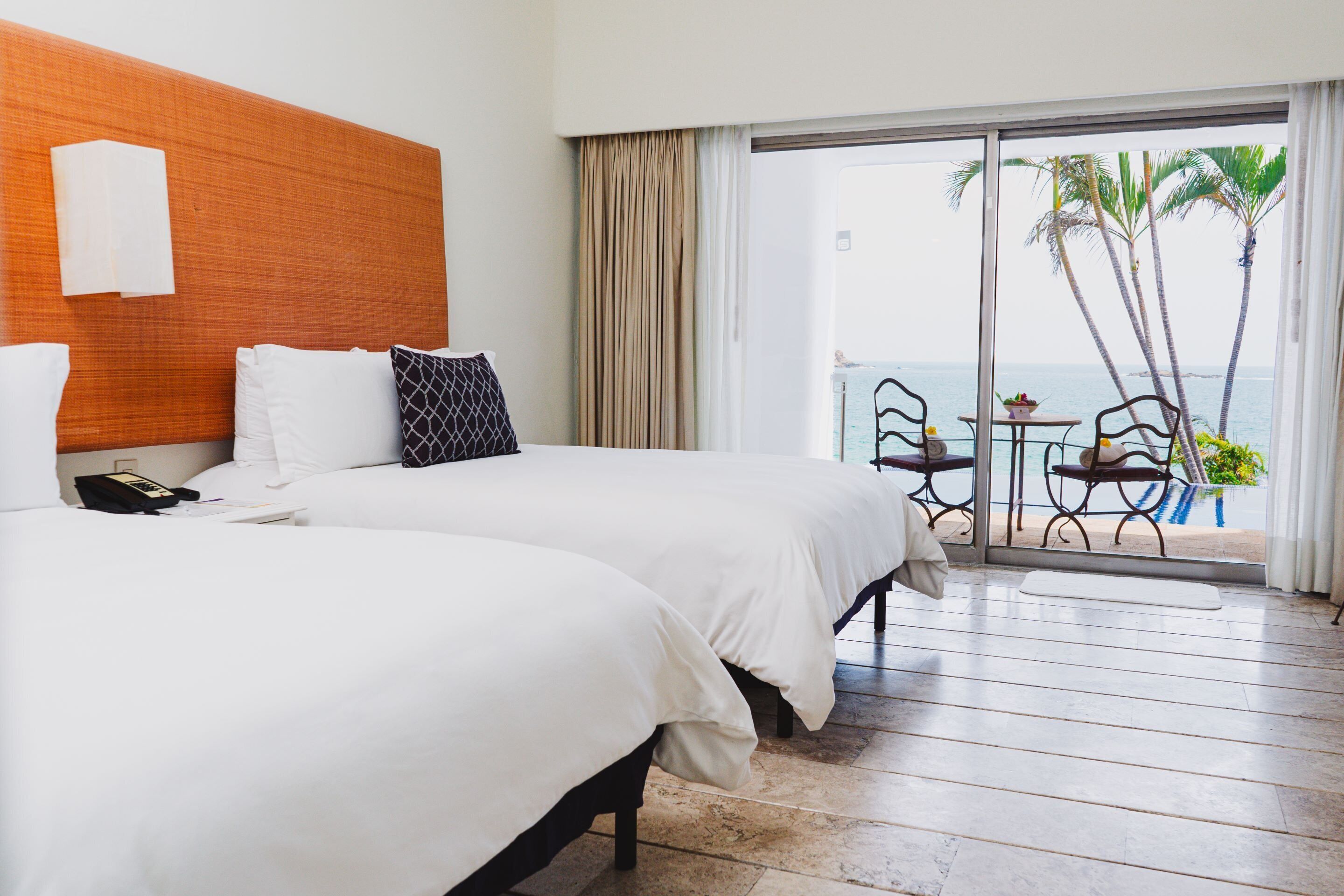 A hotel room with two beds, a wooden headboard, a balcony with a table and chairs, and a view of the ocean.
