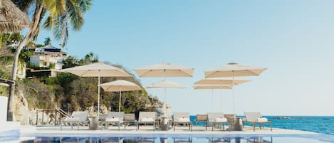 A pool area with lounge chairs and umbrellas, a clear sky, and a view of the sea.