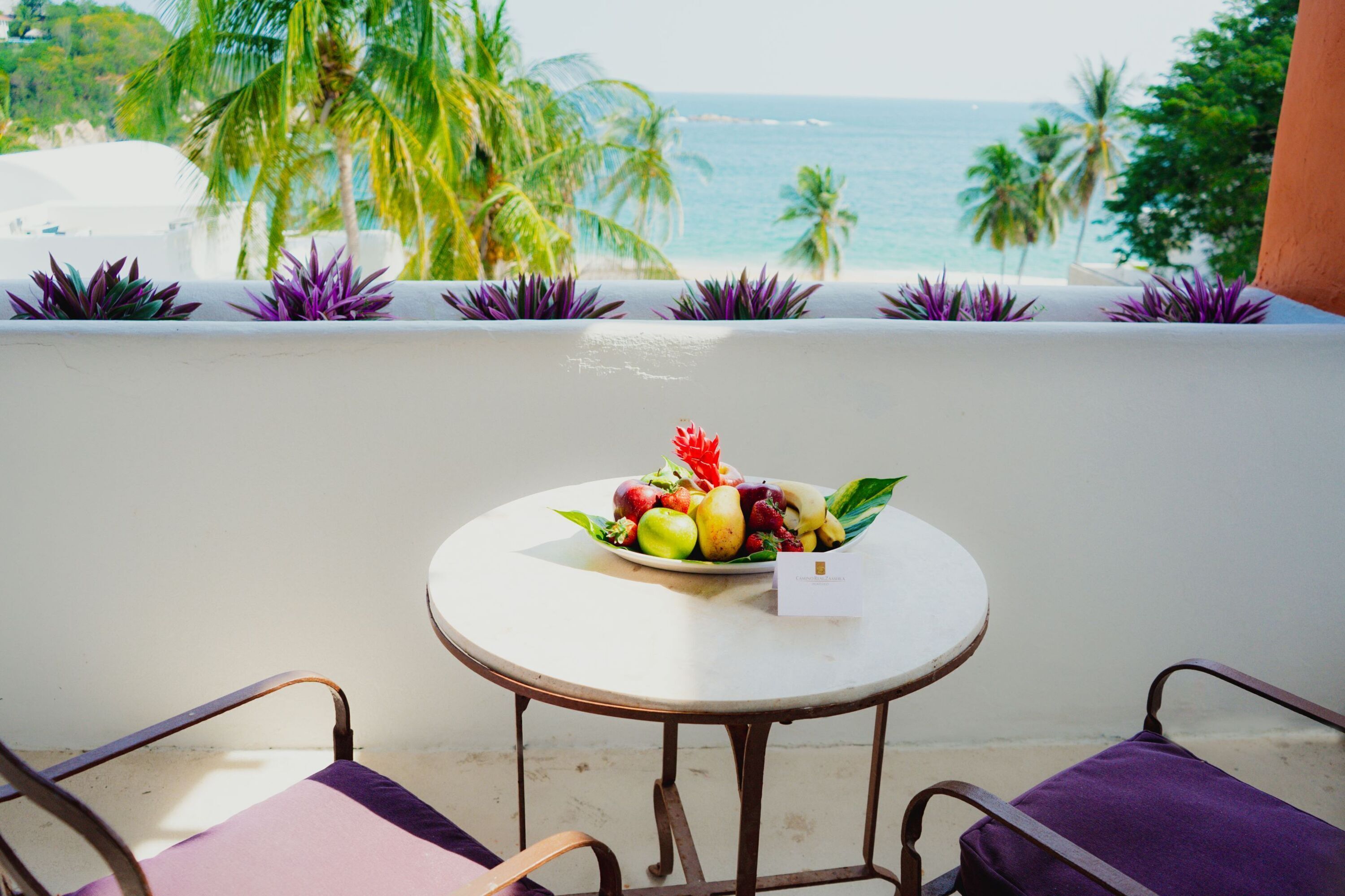A round table with a fruit bowl and a card on it, two chairs with purple cushions, and a view of the ocean and palm trees.