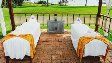 Two plush beds with white linens and yellow blankets sit on a brick-paved area, overlooking a lush lawn and the ocean.