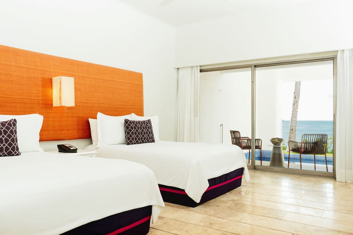 Ein Hotelzimmer mit zwei Betten, einem Holzkopfende, einem großen Fenster mit Meerblick und einem Balkon mit Sitzgelegenheit.