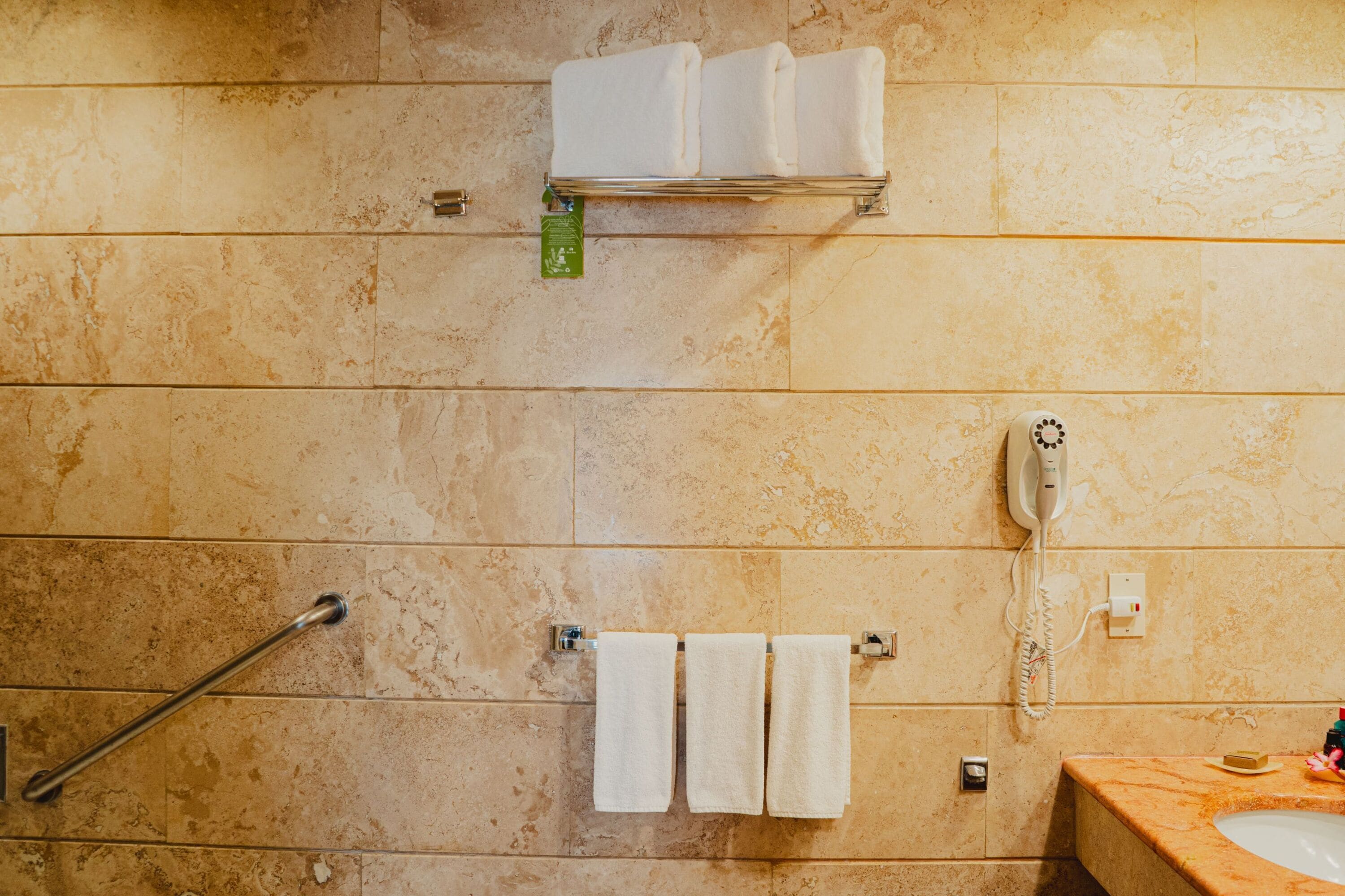 A bathroom with beige tiles, a handrail, a towel rack with two towels, a telephone, and a sink with soap.