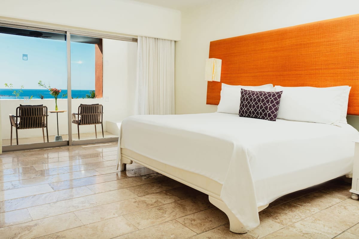 Ein Hotelzimmer mit einem großen Bett, einem Holzkopfende, einem Balkon mit Meerblick und zwei Stühlen.