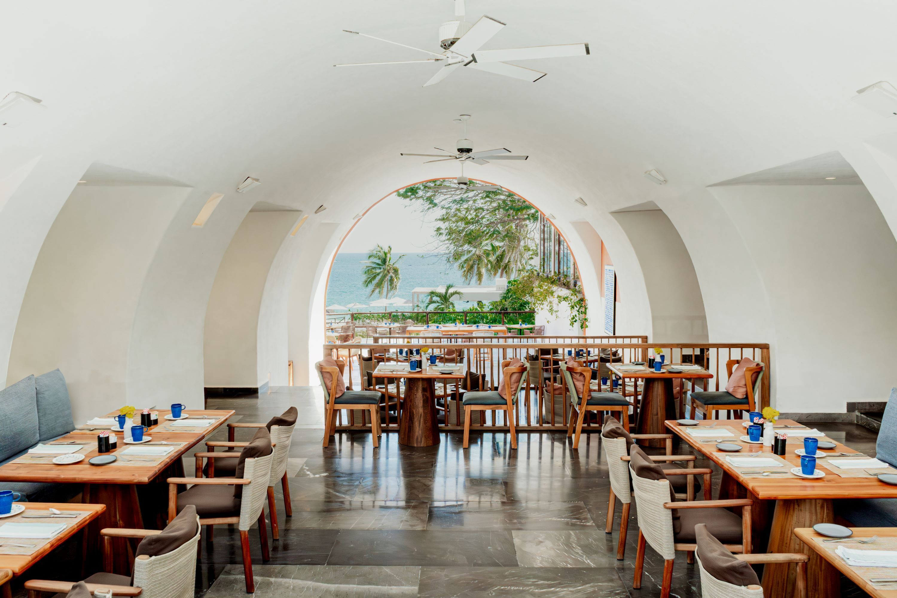 Un restaurant offrant une vue sur l’océan, avec des tables et des chaises en bois, et un ventilateur de plafond.
