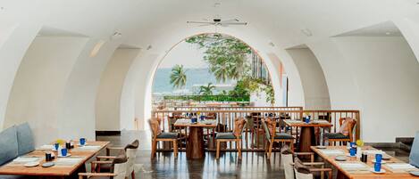 Un restaurant offrant une vue sur l’océan, avec des tables et des chaises en bois, et un ventilateur de plafond.