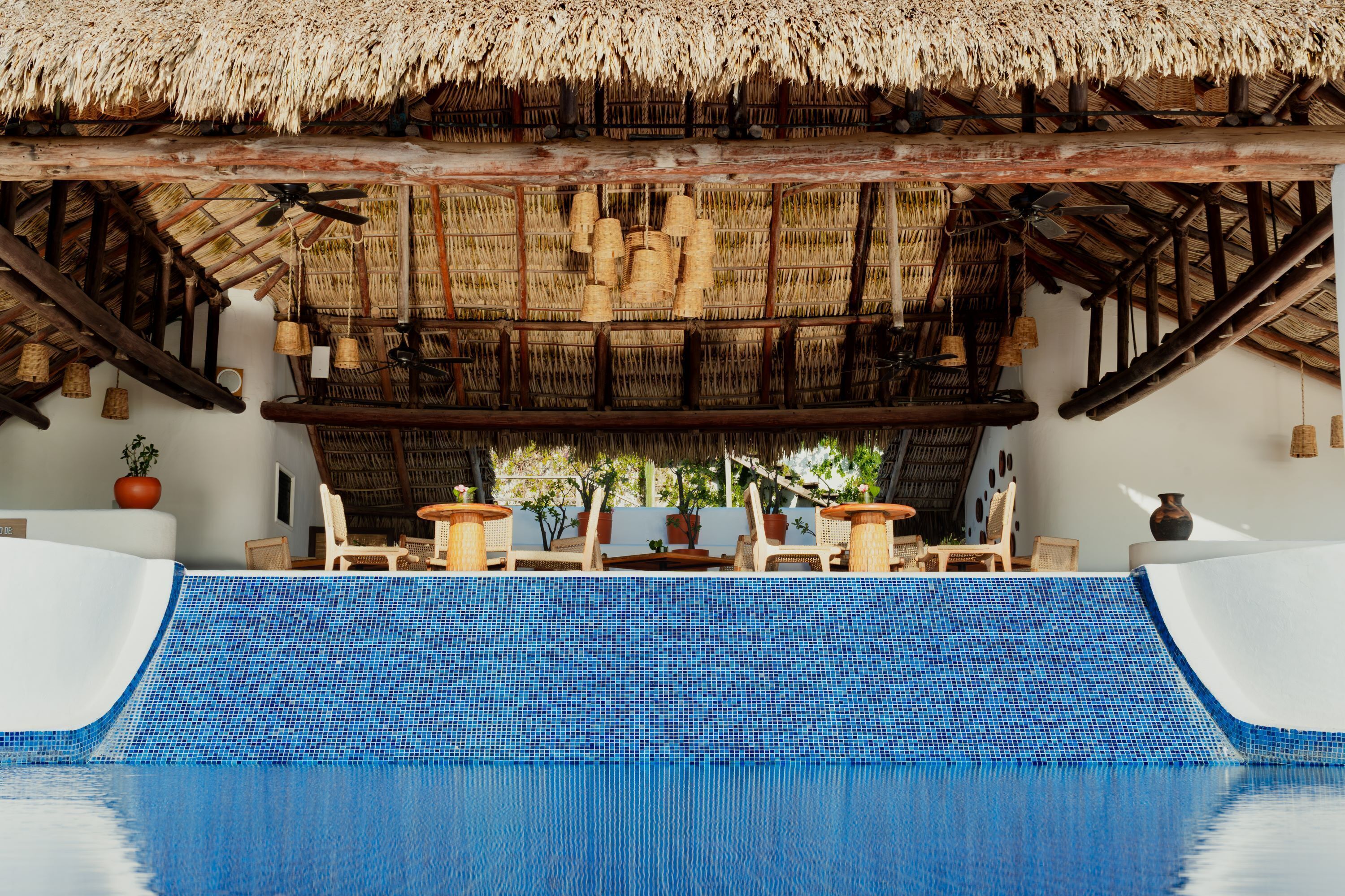 A pool area with a blue tiled floor, wooden lounge chairs, and a thatched roof structure.