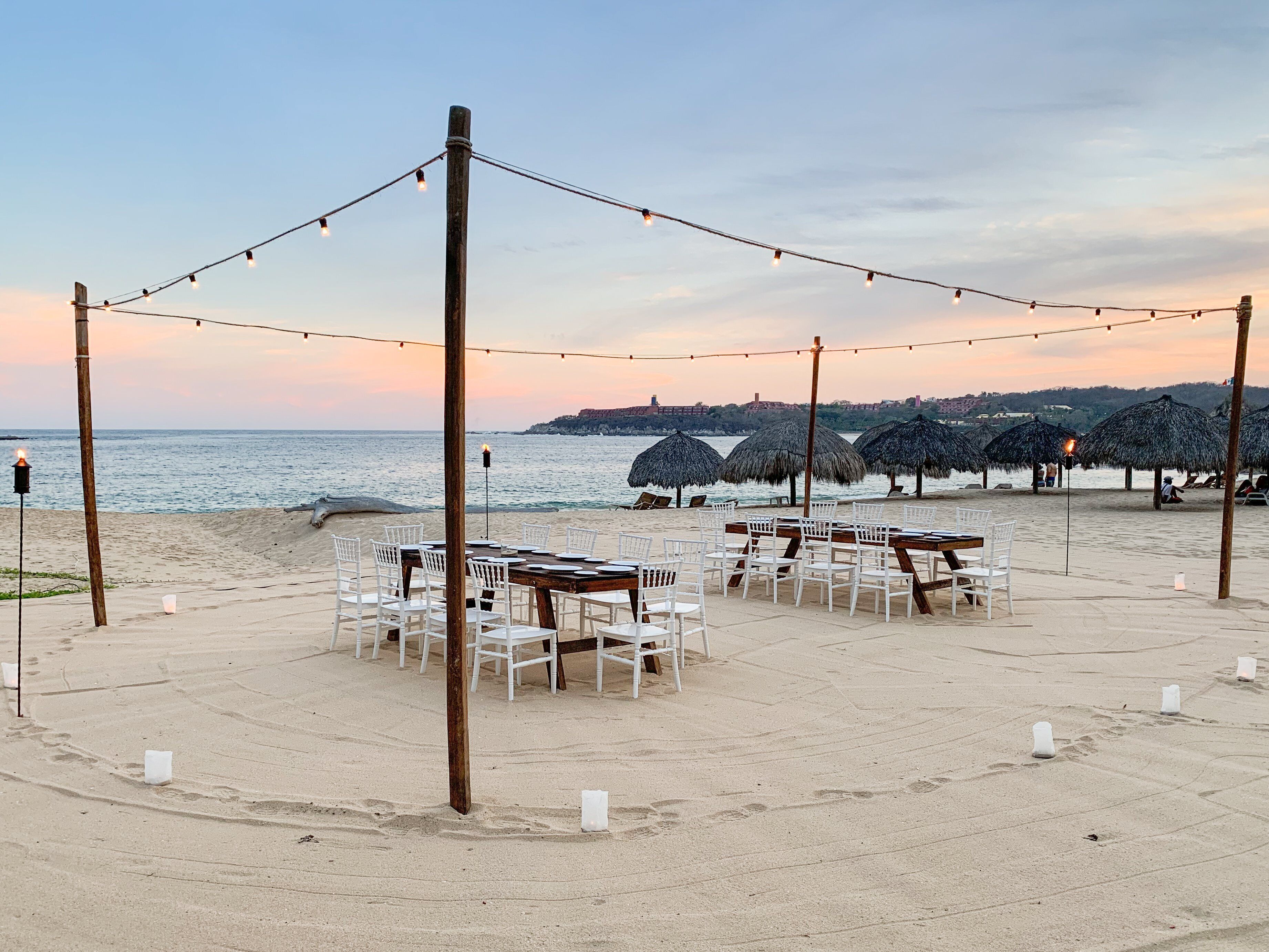 A beachside dining setup with tables and chairs, string lights, and thatched umbrellas.