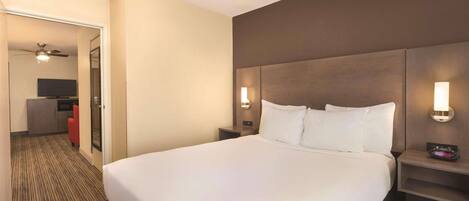 Habitación de hotel con una cama grande, una mesita de noche con lámpara, un televisor montado en la pared y un ventilador de techo.