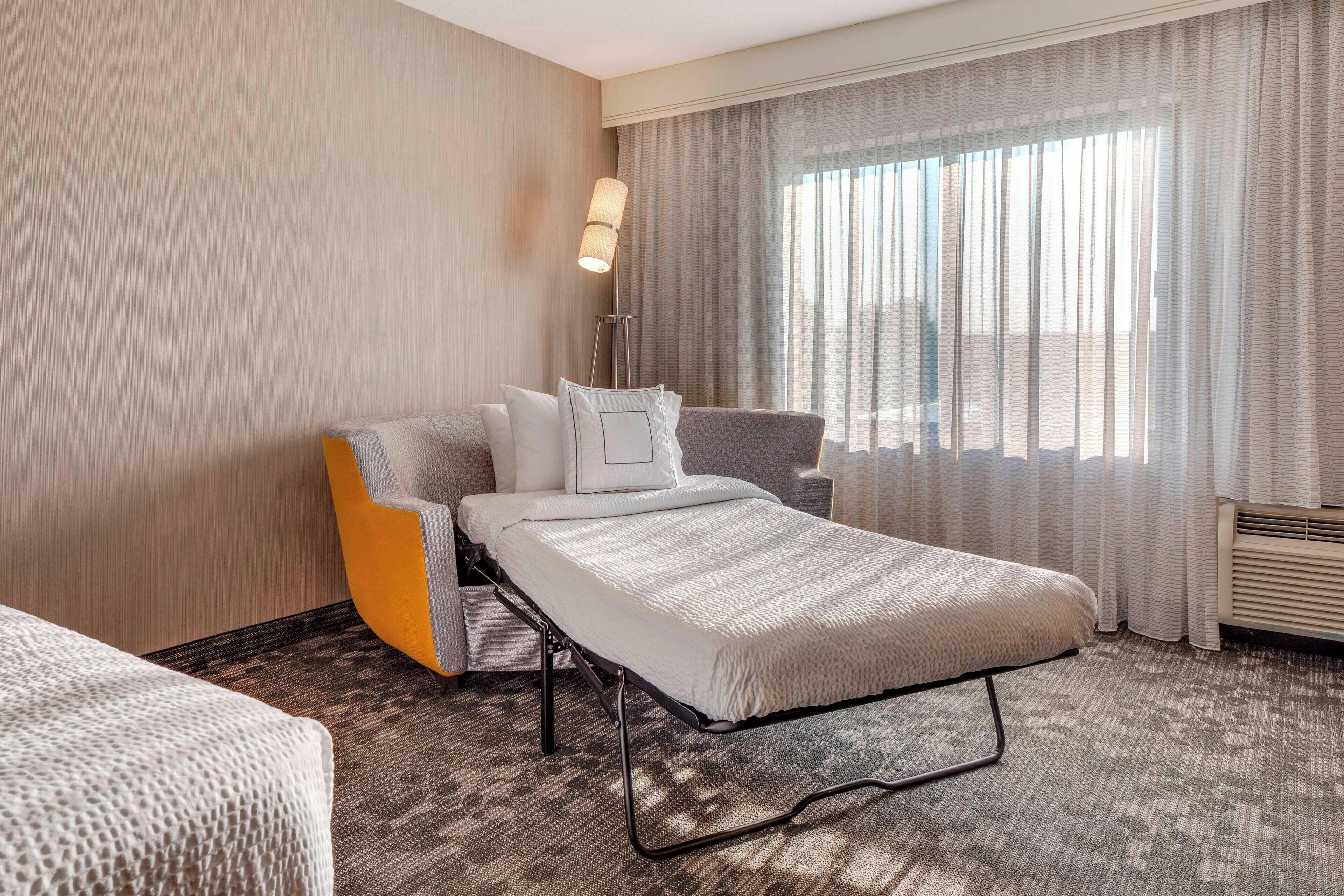 A hotel room with a single bed, a chair, a standing lamp, and a large window with sheer curtains.