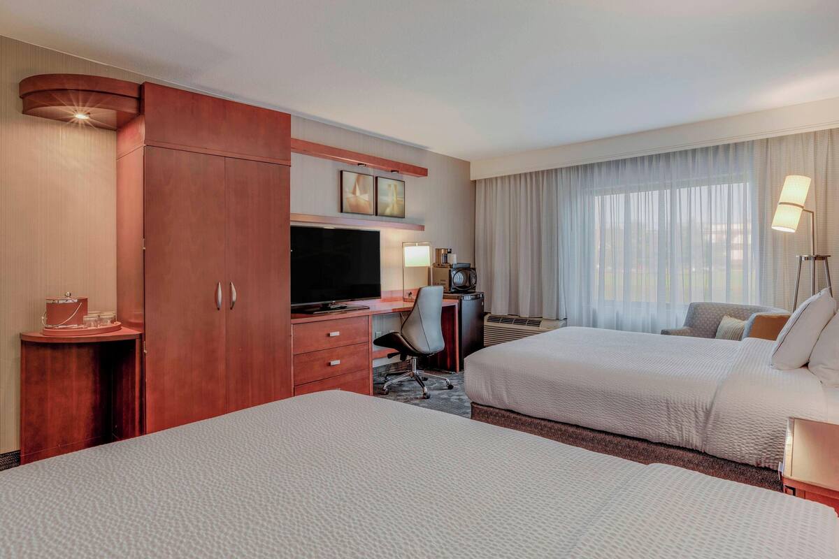 A hotel room with two beds, a desk with a TV, a minibar, and a large window with curtains.